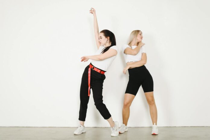Young female dancers in sportswear and sneakers making dancing movements while standing on concrete floor in studio near white wall